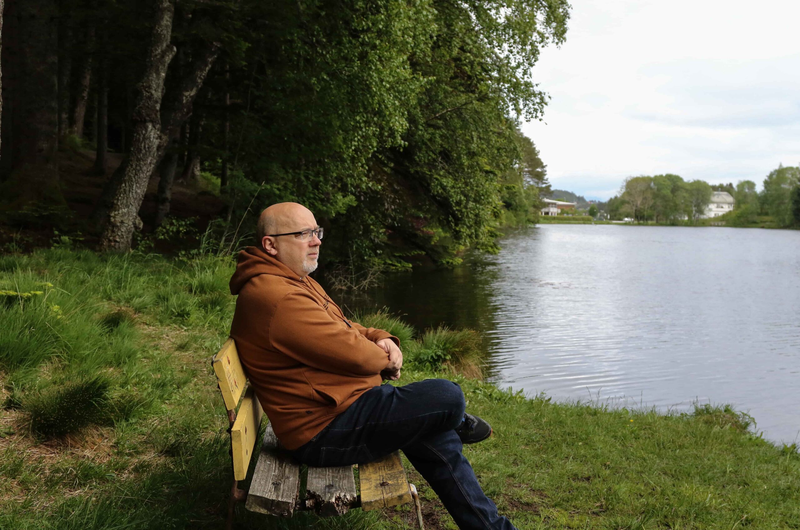 Arne sitter på en trebenk og ser utover et vann.