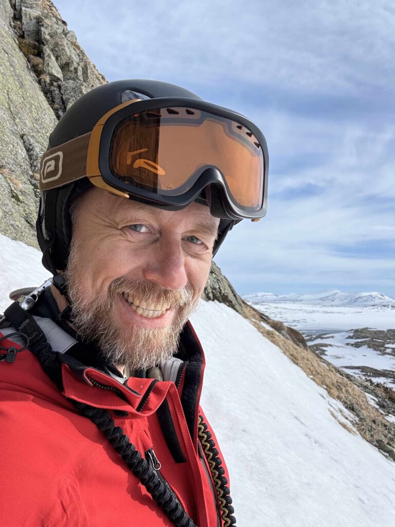 Geir Aker ser i kamera. Han har på seg alpinutstyr og det er fjell og snø i bakgrunnen. 
